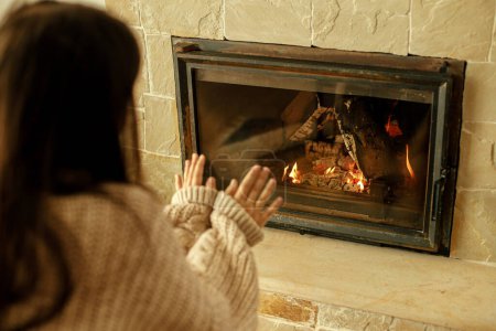 Photo for Heating house in winter with wood burning stove. Woman in cozy sweater warming up hands at fireplace in rustic room. Young stylish female sitting at fireplace in farmhouse - Royalty Free Image