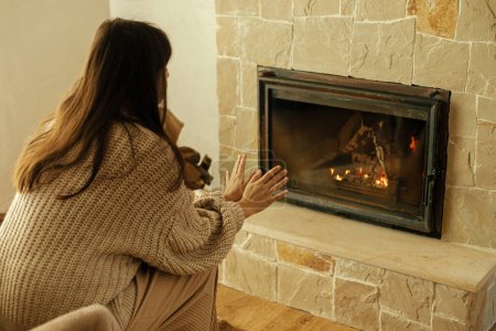 Woman in cozy sweater warming up hands at fireplace in rustic room. Young stylish  female sitting at fireplace in farmhouse. Fireplace heating in winter, alternative to gas and electricity.