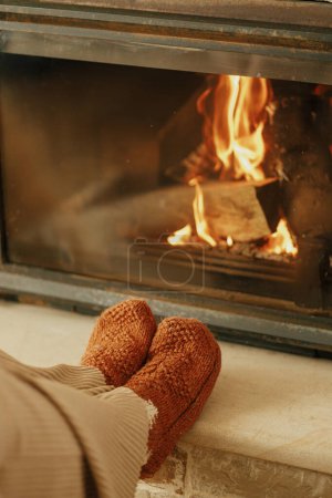 Photo for Heating house in winter with wood burning stove. Woman in cozy wool socks warming up feet at fireplace in rustic room. Young stylish female sitting at fireplace in farmhouse - Royalty Free Image
