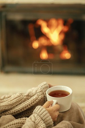 Hands in cozy sweater holding cup of warm tea on background of burning fireplace close up, autumn hygge. Heating house with wood burning stove. Relaxing and warming up at rustic fireplace