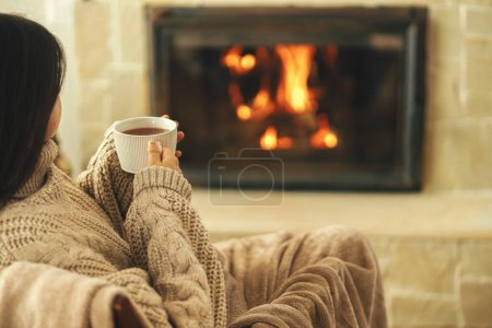 Foto de Mujer en suéter acogedor sosteniendo taza de té caliente en la chimenea en habitación rústica. Casa de calefacción en invierno con estufa de leña. Joven mujer elegante relajarse en la chimenea en la casa de campo - Imagen libre de derechos