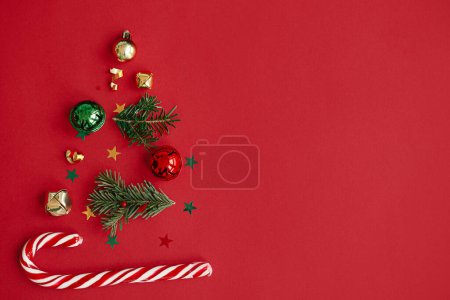 Merry Christmas! Stylish christmas tree made of fir branches, baubles, gold confetti and candy cane on red background, flat lay. Creative idea. Winter holidays, modern festive banner