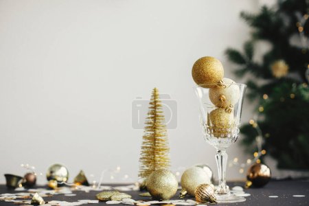 Téléchargez les photos : Merry Christmas and Happy New Year! Stylish christmas glitter baubles in champagne glass and golden decorations on dark table. Creative new year party composition. Moody holiday banner - en image libre de droit