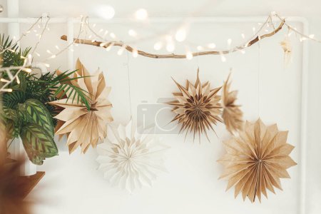 Élégantes étoiles de Noël en papier et lumières suspendues sur fond de mur blanc. Décoration festive scandinave dans la chambre boho. Papier à la main étoiles suédoises et guirlande. Temps de Noël atmosphérique