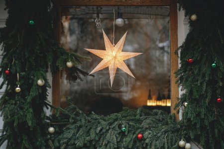 Téléchargez les photos : Élégante étoile de Noël illuminée dans les branches de fenêtre et de sapin avec des boules sur l'extérieur du bâtiment. Décor de Noël moderne dans la rue de la ville. Vacances d'hiver en Europe. Joyeux Noël - en image libre de droit