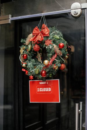 Stilvolle Weihnachtskranz rote Schleife und Kugeln und Sorry wir geschlossen Zeichen an Türen des Gebäudes. Moderne Weihnachtsdekoration in der City Street. Winterurlaub in Europa. Frohe Weihnachten
