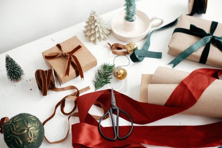 Wrapping stylish christmas gift. Modern christmas gift box, ribbons, craft paper and ornaments composition on white wood. Merry Christmas and Happy Holidays! Aesthetic winter holidays Poster 621046340
