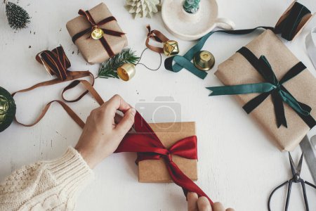 Hands wrapping stylish christmas gift flat lay. Person preparing  christmas present with red ribbon, craft paper and ornaments composition on white wood. Merry Christmas! Aesthetic winter holidays Poster 621046412
