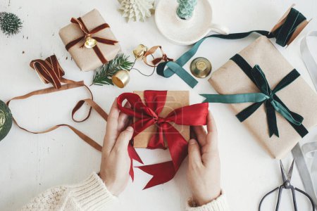 Foto de Merry Christmas! Hands wrapping stylish christmas gift flat lay. Person preparing  christmas present with red ribbon, craft paper and ornaments composition on white wood. Aesthetic winter holidays - Imagen libre de derechos
