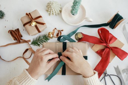 Hands wrapping stylish christmas gift flat lay. Person preparing  christmas present with green ribbon and ornaments composition on white wood. Merry Christmas! Aesthetic winter holidays Poster 621046444
