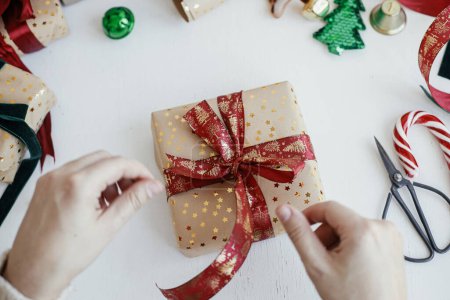 Foto de Person preparing modern gift box with red ribbon, golden wrapping paper, ornaments on white background. Merry Christmas! Hands wrapping stylish christmas gift flat lay. Atmospheric winter time - Imagen libre de derechos