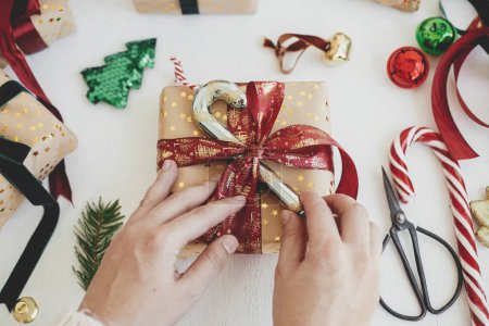 Foto de ¡Feliz Navidad! Manos decorando elegante regalo de Navidad. Persona preparando moderna caja de regalo con bastón de caramelo, cinta roja, papel de regalo, adornos sobre fondo blanco. Tiempo de invierno atmosférico - Imagen libre de derechos