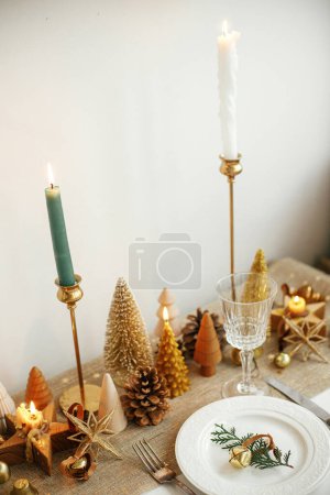 Photo for Christmas luxury table setting. Cedar branch with bell on plate, vintage cutlery, glasses, golden little christmas trees and ornaments on table. Holiday arrangement of table - Royalty Free Image