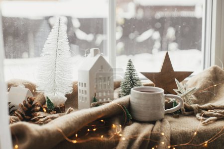 Weihnachtsstillleben, Winterhygge nach Hause. Warme Tasse Tee, Weihnachtsdekoration, Lichter, Häuschen, Stern auf kuscheliger Decke auf der Fensterbank. Gemütliches Zuhause. Atmosphärische skandinavische Stimmung