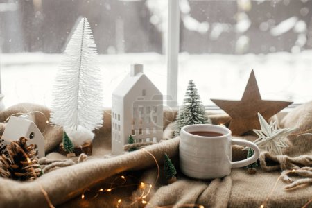 Warme Tasse Tee, Weihnachtsdekoration, Lichter, Häuschen, Stern auf kuscheliger Decke auf der Fensterbank. Winterhygge, Weihnachtsstillleben. Gemütliches Zuhause an einem verschneiten Tag. Atmosphärische skandinavische Stimmung
