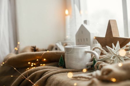 Foto de Caliente taza de té, decoraciones navideñas, luces, casita, estrella en una acogedora manta en el alféizar de la ventana. Hygge de invierno, naturaleza muerta de Navidad. Acogedora casa en un día nevado. Ambiente escandinavo atmosférico - Imagen libre de derechos