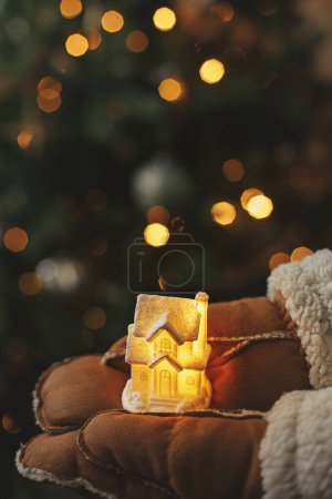 Téléchargez les photos : Mains dans les gants tenant petite maison rayonnante sur fond de lumières de sapin de Noël éclairées. Temps d'hiver magique, image atmosphérique. Confortable maison. Joyeux Noël et Joyeuses Fêtes! - en image libre de droit