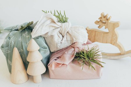 Photo for Stylish wrapped gifts in linen fabric on white rustic table with eco wooden tree, deer, fir branches. Zero waste Christmas. Furoshiki gift wrapping. Eco friendly toys. Merry Christmas! - Royalty Free Image