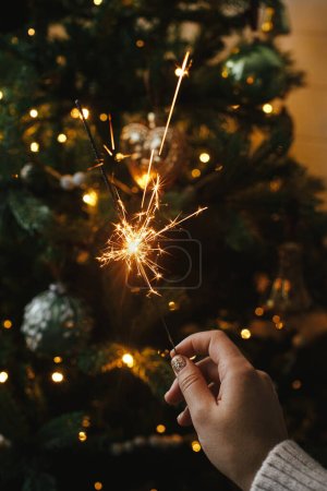 Foto de Mano sosteniendo fuegos artificiales contra las luces del árbol de Navidad en la habitación oscura. ¡Feliz Año Nuevo! ¡Feliz Navidad! Brillo ardiente en mano femenina sobre fondo de bokeh iluminación dorada. Tiempo atmosférico - Imagen libre de derechos