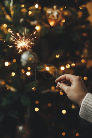 Téléchargez les photos : Main tenant un feu d'artifice contre les lumières du sapin de Noël dans la pièce sombre. Bonne année ! Joyeux Noël ! Brûlant scintillant à la main féminine sur fond de bokeh d'illumination doré. Temps atmosphérique - en image libre de droit