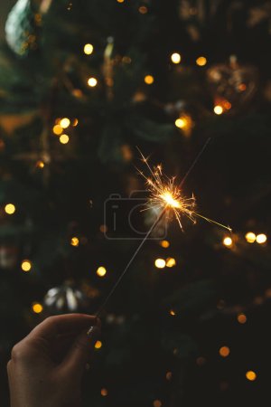 Téléchargez les photos : Bonne année ! Brûlant scintillant à la main féminine sur fond de lumières d'arbre de Noël dans la pièce sombre. Célébration atmosphérique. Main tenant feu d'artifice contre arbre décoré élégant avec éclairage - en image libre de droit