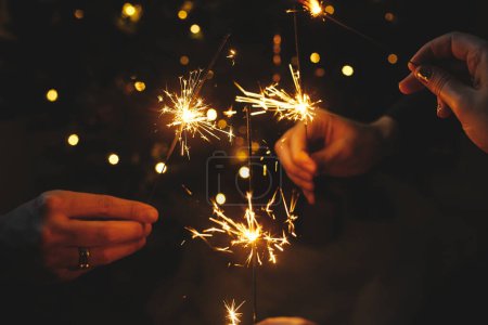 Photo for Hands holding fireworks against christmas lights in dark room. Happy New Year! Atmospheric holiday. Friends celebrating with burning sparklers in hands on background of stylish illuminated tree - Royalty Free Image
