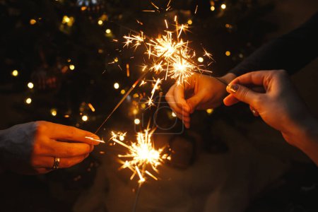 Photo for Friends celebrating with burning sparklers in hands against christmas lights in dark room. Happy New Year! Atmospheric holiday. Hands holding fireworks on background of stylish illuminated tree - Royalty Free Image