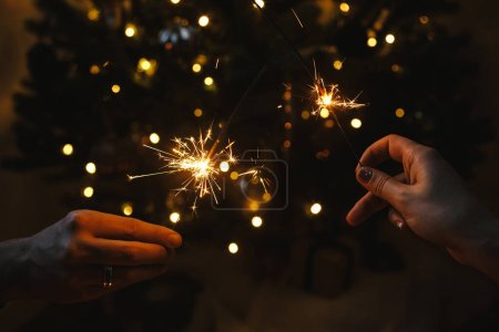 Foto de Manos sosteniendo fuegos artificiales contra las luces del árbol de navidad en la habitación oscura. ¡Feliz Año Nuevo! Pareja celebrando con bengalas ardientes en las manos sobre el fondo del árbol adornado de estilo con la iluminación. - Imagen libre de derechos
