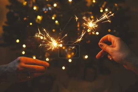Photo for Happy New Year! Couple celebrating with burning sparklers in hands on background of stylish decorated tree with illumination. Hands holding firework against christmas tree lights in dark room - Royalty Free Image