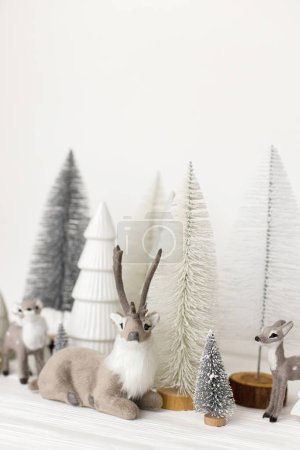 Foto de ¡Feliz Navidad! Pequeños árboles de Navidad con estilo y juguetes de renos sobre fondo blanco. Escena navideña festiva, bosque nevado en miniatura con ciervos. Decoraciones modernas, banner de vacaciones - Imagen libre de derechos