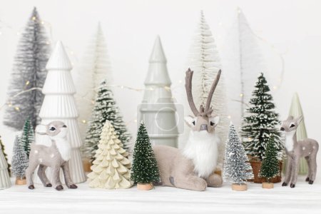 Foto de ¡Feliz Navidad! Pequeños árboles de Navidad con estilo y juguetes de renos sobre fondo blanco. Escena navideña festiva, bosque nevado en miniatura con ciervos. Decoraciones modernas, banner de vacaciones - Imagen libre de derechos