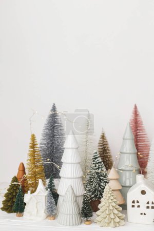 Photo for Merry Christmas! Stylish little Christmas trees and houses decorations on white table. Modern christmas scene, miniature cozy snowy village. Winter holiday banner, scandinavian decor - Royalty Free Image