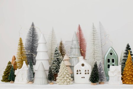 Foto de ¡Feliz Navidad! Pequeños árboles de Navidad con estilo y casas decoraciones en mesa blanca. Escena navideña moderna, pequeño y acogedor pueblo nevado. Banner de vacaciones de invierno, decoración escandinava - Imagen libre de derechos