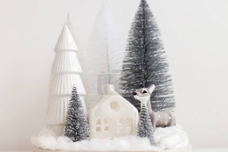 Téléchargez les photos : Petits arbres de Noël élégants et jouet de renne sur table blanche. Scène de Noël festive, forêt enneigée miniature avec des cerfs mignons. Joyeux Noël ! Cadre de table moderne, décorations monochromes - en image libre de droit