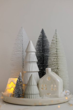 Foto de Hygge de invierno, acogedora escena mágica de Navidad, pueblo nevado en miniatura con luces. Pequeños árboles de Navidad con estilo y la casa en la mesa blanca. ¡Feliz Navidad! Decoración moderna de vacaciones, tiempo atmosférico - Imagen libre de derechos