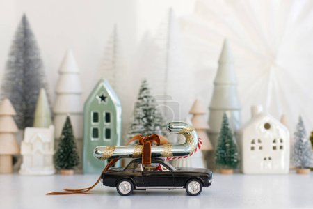 Téléchargez les photos : Les vacances arrivent ! Élégante petite voiture portant une canne à bonbons sur fond de village enneigé miniature de Noël. Joyeux Noël ! Scène d'hiver festive sur table blanche, bannière de Noël - en image libre de droit