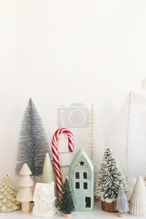 Photo for Merry Christmas and Happy Holidays! Stylish little Christmas trees, houses and candy cane on white table. Modern christmas scene, miniature festive snowy village. Winter banner, scandinavian decor - Royalty Free Image