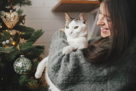 Photo for Woman in cozy sweater hugging cute cat near stylish christmas tree with vintage baubles. Pet and winter holidays. Adorable kitty and happy woman cuddling in festive room. Merry Christmas! - Royalty Free Image