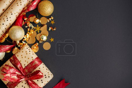 Foto de Elegante regalo de Navidad con cinta roja, papel de regalo, confeti dorado y adornos sobre fondo negro. Moderno regalo de Navidad plana con espacio para el texto. ¡Feliz Navidad! Banner de vacaciones - Imagen libre de derechos