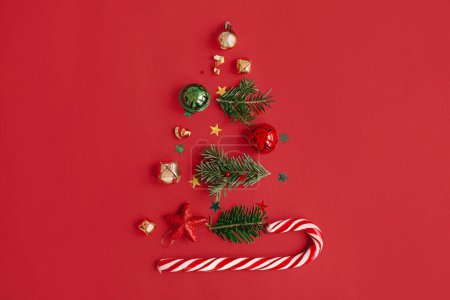 Téléchargez les photos : Arbre de Noël, concept vacances d'hiver. Élégant arbre de Noël composé de branches de sapin, de boules, de confettis d'or et de canne à sucre sur fond rouge, plat. Idée créative. Bannière festive moderne - en image libre de droit