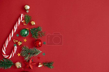 Téléchargez les photos : Arbre de Noël, concept vacances d'hiver. Élégant arbre de Noël composé de branches de sapin, de boules, de confettis d'or et de canne à sucre sur fond rouge, plat. Idée créative. Bannière festive moderne - en image libre de droit