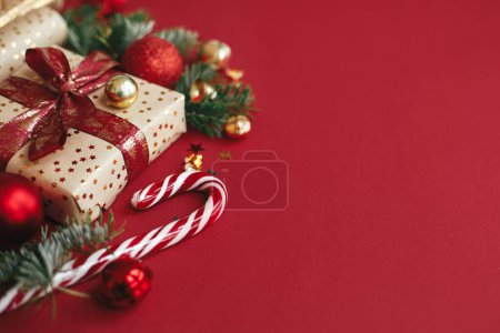 Foto de Composición navideña moderna. Elegante regalo de Navidad, papel de regalo, ramas de abeto, bastón de caramelo y adornos sobre fondo rojo, espacio para el texto. Feliz Navidad y Felices Fiestas! Banner festivo - Imagen libre de derechos