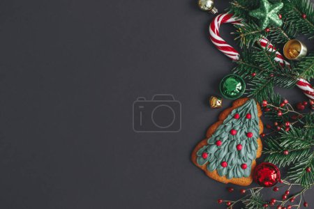 Stylish christmas cookie, tree branches, candy cane, festive decorations border on black background. Modern christmas flat lay, space for text. Season's greetings card. Merry Christmas!