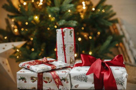 Foto de Elegantes regalos de Navidad en el árbol de Navidad con luces doradas bokeh. Envuelto regalos de Navidad con cinta roja bajo el árbol decorado en la habitación. Víspera atmosférica. Banner navideño. Feliz Navidad.! - Imagen libre de derechos
