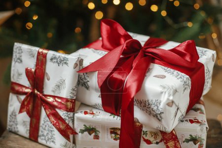 Téléchargez les photos : Cadeaux de Noël élégants à l'arbre de Noël avec des lumières. Joyeux Noël et Joyeuses Fêtes ! Cadeaux de Noël enveloppés avec ruban rouge sous l'arbre décoré dans une pièce humide. Bannière atmosphérique - en image libre de droit