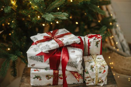 Téléchargez les photos : Cadeaux de Noël élégants à l'arbre de Noël avec des lumières. Joyeux Noël et Joyeuses Fêtes ! Cadeaux de Noël enveloppés avec ruban rouge sous l'arbre décoré dans une pièce humide. Bannière atmosphérique - en image libre de droit