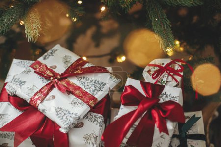 Foto de Feliz Navidad y Felices Fiestas! Elegantes regalos de Navidad en el árbol de Navidad con luces doradas bokeh. Envuelto regalos de Navidad con cinta roja bajo el árbol decorado en la habitación. Banner atmosférico - Imagen libre de derechos