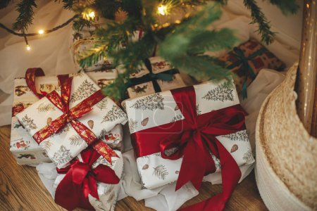 Foto de Elegantes regalos de Navidad en el árbol de Navidad con luces doradas bokeh. Envuelto regalos de Navidad con cinta roja bajo el árbol decorado en la habitación. Víspera atmosférica. Banner navideño. Feliz Navidad.! - Imagen libre de derechos