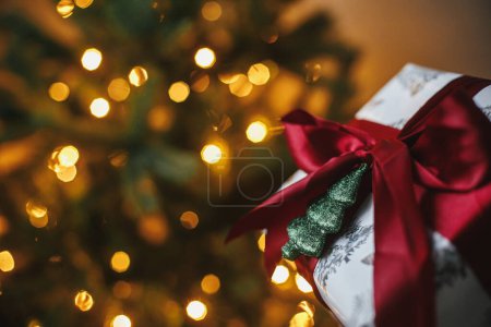 Foto de ¡Feliz Navidad! Elegante regalo de Navidad en el árbol de Navidad con luces doradas. Envuelto regalo de Navidad con lazo rojo y adorno de árbol brillante en la sala de la noche. Banner atmosférico, espacio para texto - Imagen libre de derechos