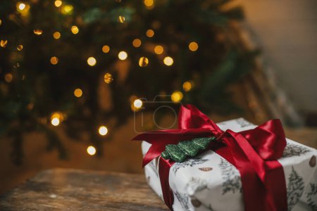 Foto de Elegante regalo de Navidad en el árbol de Navidad con luces doradas. Envuelto regalo de Navidad con lazo rojo y adorno de árbol brillante en la sala de la noche. ¡Feliz Navidad! Banner atmosférico, espacio para texto - Imagen libre de derechos